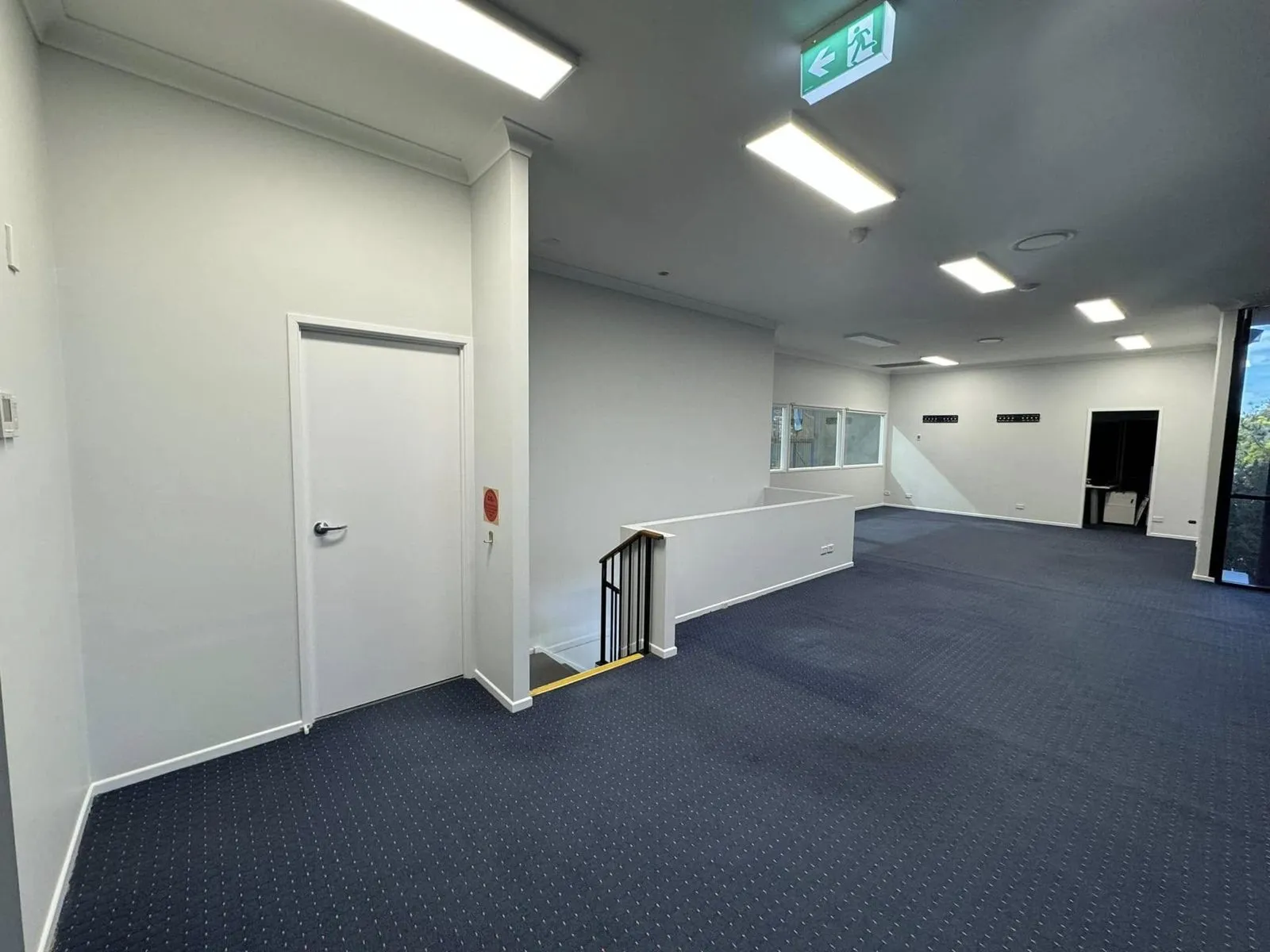 Upper office floor with white walls and ceiling, a white door, a stairwell and dark carpet